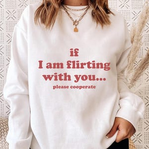 Może przedstawiać: Biała bluza z napisem "if I am flirting with you... please cooperate" w kolorze czerwonobrązowym. Bluza ma okrągły dekolt i długie rękawy. Tło jest w wzór w kolorze złamanej bieli.
