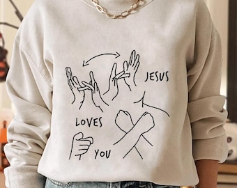 May include: Beige sweatshirt with a black graphic of hands in sign language spelling "Jesus Loves You." The design includes arrows and the words "LOVES" and "YOU."