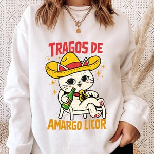 May include: White sweatshirt featuring a cartoon cat wearing a yellow sombrero and holding a bottle. The text "TRAGOS DE AMARGO LICOR" is printed in red and yellow. The cat is sitting on a chair with a green bottle in its paw.