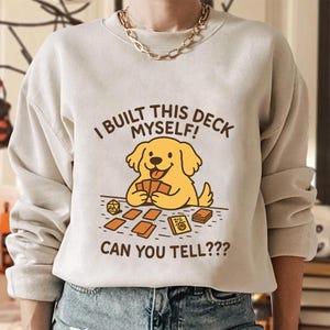 Może przedstawiać: Beżowa bluza z nadrukiem żółtego psa z kreskówki trzymającego karty, z brązowym napisem "I BUILT THIS DECK MYSELF! CAN YOU TELL???". Pies siedzi na stole z kartami i kośćmi do gry.
