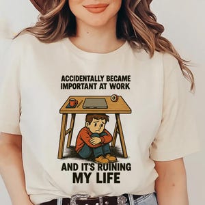 May include: A cream-coloured t-shirt featuring the text "ACCIDENTALLY BECAME IMPORTANT AT WORK AND IT'S RUINING MY LIFE." The graphic shows a person under a desk with a laptop, coffee cup, and tape.