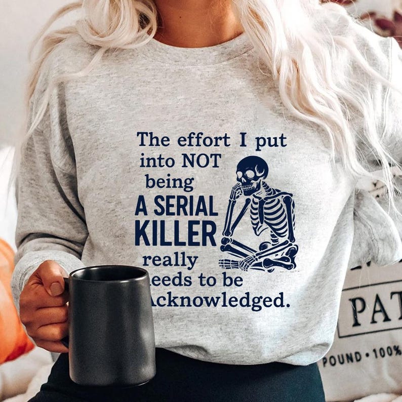 May include: A light grey sweatshirt featuring the text "The effort I put into NOT being A SERIAL KILLER really needs to be acknowledged." A blue skeleton graphic is also on the sweatshirt. A black mug is held.