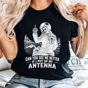 以下が含まれることがあります： バイクに乗った骸骨のバイカーが中指を立てている黒いTシャツ。バイカーはヘルメットと「RIDE FREE LIVE FREE」の文字が入ったジャケットを着ています。Tシャツには「CAN YOU SEE ME BETTER IF I PUT UP MY ANTENNA」の文字も入っています。