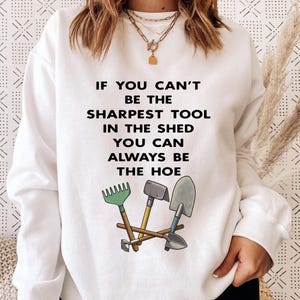 Può includere: Felpa bianca con la scritta "IF YOU CAN'T BE THE SHARPEST TOOL IN THE SHED YOU CAN ALWAYS BE THE HOE." Il design include illustrazioni di un rastrello, un martello e una pala.