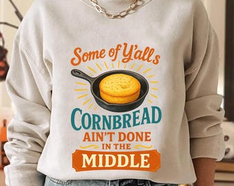 May include: Beige sweatshirt with the text "Some of Y'alls Cornbread Ain't Done in the Middle." The graphic features a cornbread in a skillet with yellow rays. The sweatshirt is a casual, comfortable style.