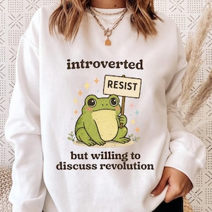 May include: White sweatshirt featuring the text "introverted resist but willing to discuss revolution." A cartoon frog, holding a "resist" sign, is the central image. The frog is green and yellow, surrounded by stars and plants.