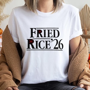May include: White t-shirt with the text "FRIED RICE '26" in a bold, black font. The word "FRIED" is above "RICE '26". The letters "R" and "E" in "FRIED" and "RICE" are in red. The shirt is worn by a person.