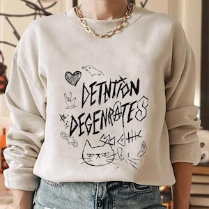 May include: A beige sweatshirt with black graphic doodles, including a heart, bird, and cat face. The words "DETENTION DEGENERATES" are written in a bold, graffiti-style font. The sweatshirt has long sleeves and a crew neck.