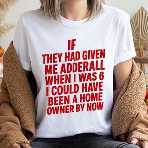 Può includere: T-shirt bianco con testo rosso: "IF THEY HAD GIVEN ME ADDERALL WHEN I WAS 6 I COULD HAVE BEEN A HOME OWNER BY NOW." La maglietta è indossata con una giacca marrone.