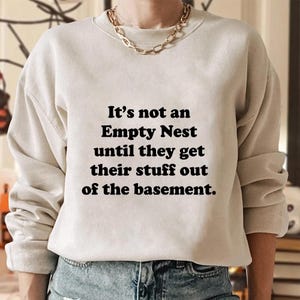Op de afbeelding: Een beige sweatshirt met de tekst "It's not an Empty Nest until they get their stuff out of the basement." De top met lange mouwen wordt gecombineerd met een blauwe jeans. Een gouden ketting is zichtbaar.