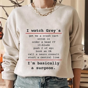 May include: A cream-coloured sweatshirt featuring the text "I watch Grey's" and a list of medical actions, concluding with "I'm basically a surgeon." The text is black, with red lines.