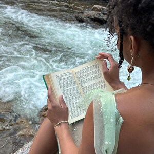 a pair of earrings made from hemlock pinecones with green glass teardrop beads modeled by a woman reading a book in the forest next to a creek