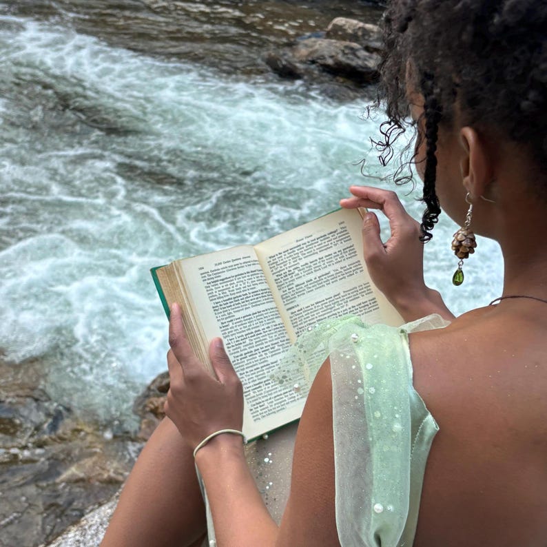 a pair of earrings made from hemlock pinecones with green glass teardrop beads modeled by a woman reading a book in the forest next to a creek