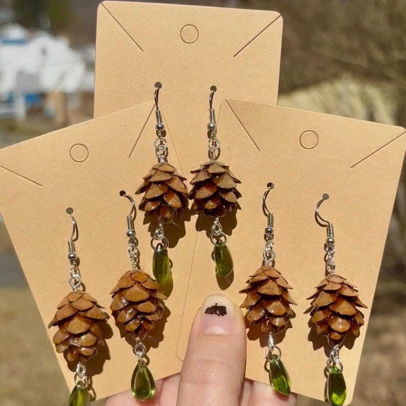 Puede incluir: Pendientes hechos a mano con pi&ntilde;as en miniatura, ganchos plateados y cuentas verdes en forma de l&aacute;grima. Los pendientes se exhiben en tarjetas de exhibici&oacute;n de pendientes de cartulina marr&oacute;n. Las pi&ntilde;as son marrones y tienen un aspecto natural.