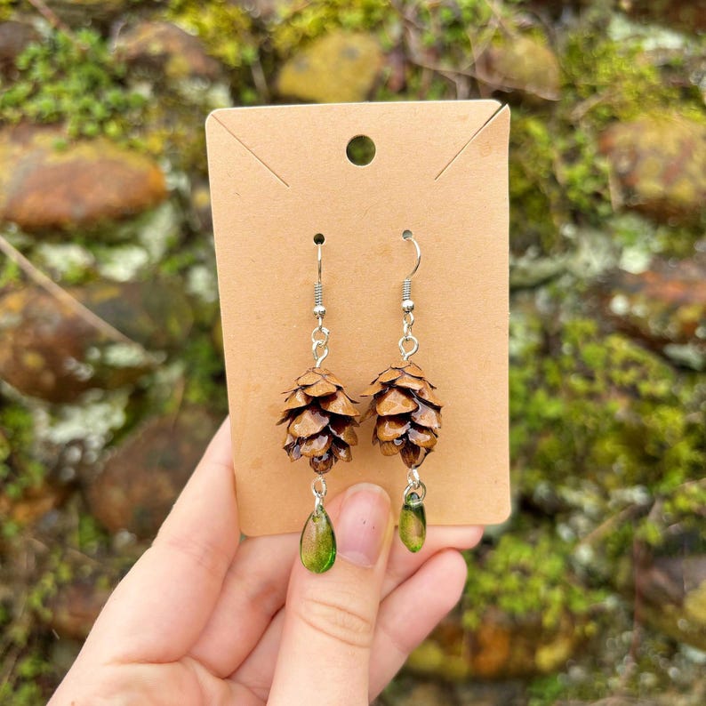 a pair of earrings made from hemlock pinecones with green glass teardrop beads on earring hooks held in front of a stone wall