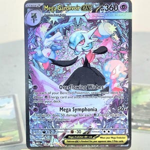 Może przedstawiać: Karta Pokémon Mega Gardevoir EX, z kolorową ilustracją postaci Gardevoir. Karta zawiera tekst "Overflowing Wishes" i "Mega Symphonia". Karta jest prezentowana na małym drewnianym sztaludze.