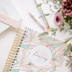 May include: A spiral-bound planner with a geometric design in pastel pink, teal, and gold. The planner is labeled "The Intentional Living Planner" and is accompanied by matching pens and a notebook. Floral accents add a touch of elegance.