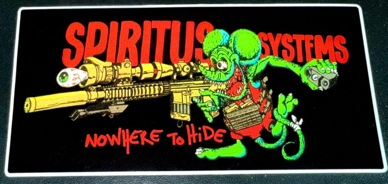 Op de afbeelding: Zwarte rechthoekige sticker met de woorden "SPIRITUS SYSTEMS" in het rood. Een cartoon groene rat met een geweer en een oog in de scope is afgebeeld. De tekst "NOWHERE TO HIDE" staat eronder.