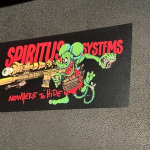 Op de afbeelding: Zwarte sticker met een cartoonrat die een geweer vasthoudt. De tekst "SPIRITUS SYSTEMS" staat in het rood, en "NOWHERE TO HIDE" staat ook in het rood. De rat is groen, rood en bruin. De sticker is rechthoekig.