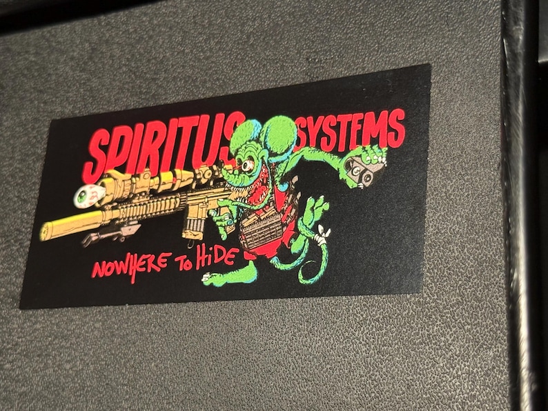 Op de afbeelding: Zwarte sticker met een cartoonrat die een geweer vasthoudt. De tekst "SPIRITUS SYSTEMS" staat in het rood, en "NOWHERE TO HIDE" staat ook in het rood. De rat is groen, rood en bruin. De sticker is rechthoekig.
