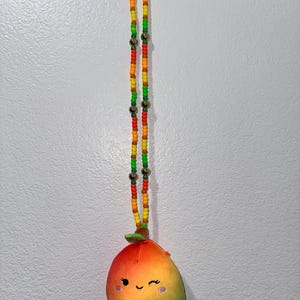 May include: A beaded necklace with a plush mango charm. The necklace features a colorful pattern of red, orange, yellow, and green beads, with gold-colored accents. The mango charm is a vibrant mix of red, orange, and green, with a smiling face.