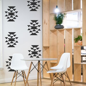 May include: A white table with two white chairs sits in a room with a white wall featuring black geometric designs. The wall is next to a wooden shelving unit.