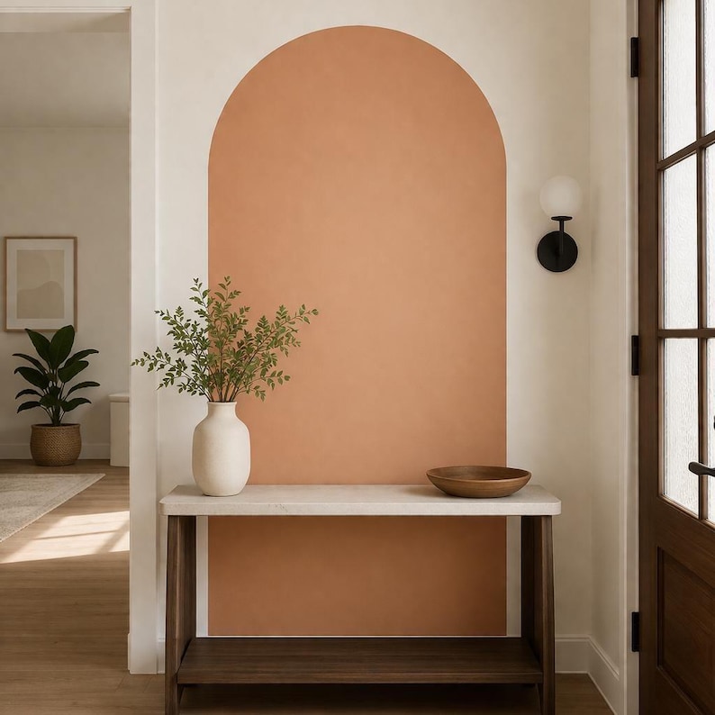 Puede incluir: Una mesa consola con tablero blanco y estructura de madera oscura se apoya contra una pared con un gran arco pintado en color terracota. Sobre la mesa hay un jarr&oacute;n de cer&aacute;mica blanca con ramas verdes y un cuenco de madera.