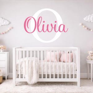 Puede incluir: Una cuna blanca con una manta blanca y esponjosa y almohadas rosas está frente a una pared con el nombre "Olivia" en escritura rosa dentro de una "O" blanca. La guardería está decorada con acentos de nubes y pompones.