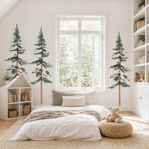 May include: A bedroom scene with a white bed, a window, and three watercolor-style pine tree wall decals. The room has a neutral color palette with a beige rug, a woven ottoman, and a white bookshelf. A teddy bear sits on the ottoman.