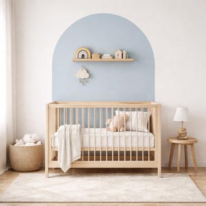 Puede incluir: Una cuna de madera clara con un colch&oacute;n blanco, una manta y un oso de peluche se encuentra sobre una alfombra crema. Una peque&ntilde;a mesita de noche de madera con una l&aacute;mpara est&aacute; al lado de la cuna. Una estanter&iacute;a con un arco&iacute;ris, libros y una casa est&aacute; montada en una pared azul.