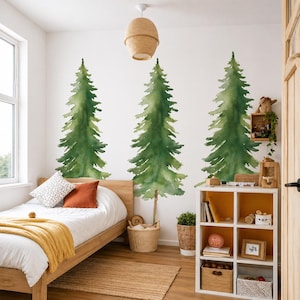 May include: A bedroom scene featuring three large green watercolor-style pine tree decals on a white wall. The room includes a wooden bed with orange and white pillows, a yellow blanket, and a white storage unit with decorative items.