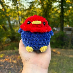 May include: A handmade, plush bird in vibrant colors. The bird features a red head, a blue body, and yellow feet. It has small black eyes and a yellow beak. The bird is made of soft, textured yarn, perfect for a child's toy or a decorative item.