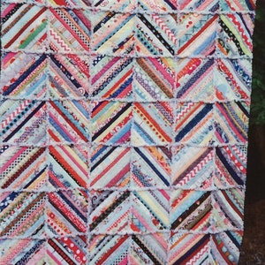 Pode incluir: Uma colcha de retalhos colorida com um padrão chevron. A colcha é feita de tiras de tecido em várias cores e padrões, incluindo vermelho, azul, rosa, amarelo e verde. A colcha é franjada e tem um forro branco.