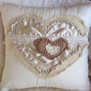 May include: A white pillow with a heart-shaped design made of different fabrics, including lace, satin, and burlap. The heart is layered and has a brown knitted heart in the center.