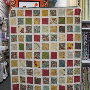 May include: A patchwork quilt featuring a range of colourful fabrics in squares. The quilt is made up of different patterns and colours, including red, green, yellow, and blue. The quilt is hanging on a wall in a shop.
