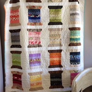 May include: A white quilt with a patchwork design of colourful spools of thread. Each spool is made up of different fabric scraps, creating a unique and textured look. The quilt is hanging on a wall, with a white wooden shelf below it.