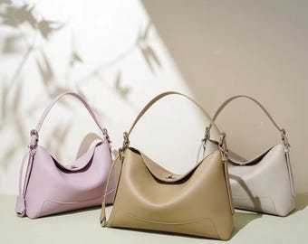 May include: Three leather handbags in different colours: light purple, beige, and light grey. The handbags have a simple, elegant design with a single strap and a small, silver clasp.