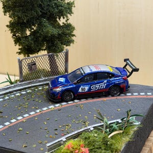 May include: A blue Subaru STI model car with racing decals, including the number 90, sits on a miniature asphalt road with a painted white line. The scene includes a small tree, a fence, and patches of greenery, creating a diorama.
