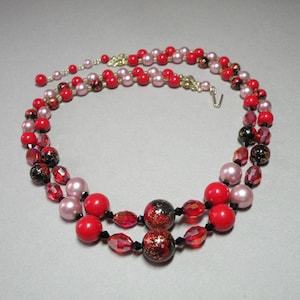 May include: A double strand necklace featuring red, pink and black beads. The beads are round and have a glossy finish. The necklace is made from glass beads.