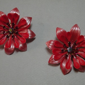 May include: A pair of red flower-shaped earrings with jeweled centres. The earrings are made of plastic and have a shiny finish.
