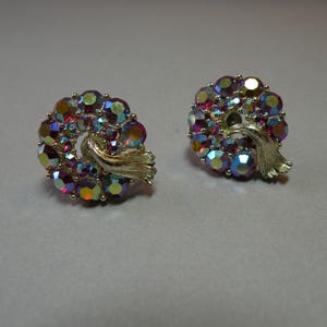 May include: A pair of iridescent earrings with a gold tone setting. The earrings are shaped like a swirl and are adorned with a cluster of colourful rhinestones.