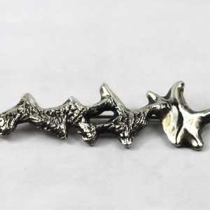 May include: A silver brooch with a textured, organic design resembling a branch or coral. The brooch is abstract and modern in style.