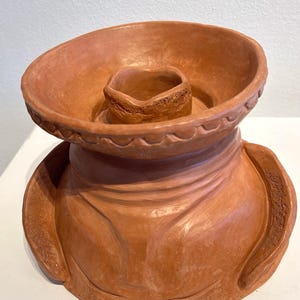 May include: A terracotta-colored ceramic sculpture with a sombrero-like design. The piece features a wide, shallow brim and a central, raised cylindrical element. The sculpture has a textured surface and is likely handmade.