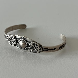Puede incluir: Una pulsera de plata con diseños detallados de inspiración nativa americana. La pulsera presenta patrones simétricos, incluyendo motivos estilizados de pájaros o plumas y símbolos de flechas. El elemento central del diseño es una cuenta circular en relieve.