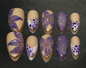 Può includere: Un set di dieci unghie finte con vari design. Alcune unghie presentano una base beige con accenti floreali viola, mentre altre hanno un design a pois viola o un colore viola pieno con una superficie strutturata. Le unghie sono di forma ovale.