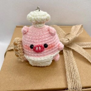 May include: A crocheted piglet in a chef's hat and apron, holding a bread roll. The piglet is pink with black eyes and a red snout. The keychain is on a brown gift box tied with a burlap ribbon.