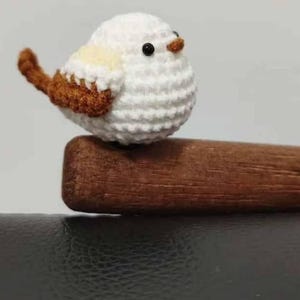 May include: A handmade, crocheted bird figurine. The bird is primarily white with brown and yellow accents on its wings and tail. It has a small, dark beak and is perched on a wooden dowel. The bird is a charming decorative item.