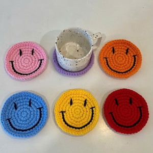May include: Six colourful, crocheted coasters, each with a smiling face design. The coasters are in pink, orange, blue, yellow, and red. A white speckled mug rests on a purple coaster. The coasters are designed to protect surfaces.