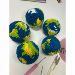 May include: Five handmade crochet balls depicting the Earth. Each ball features blue oceans, green and yellow continents, and white polar ice caps. A crochet hook and other crafting tools are visible in the foreground.