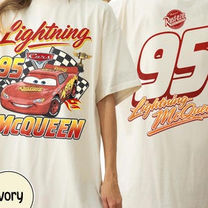 May include: Ivory t-shirt featuring Lightning McQueen from the Cars film. The front graphic includes the number 95, a chequered flag, and the words "Lightning McQueen" in red and yellow. The back displays the number 95 and "Lightning McQueen" in red and orange.